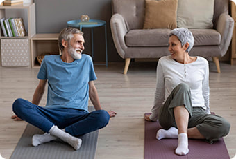 Dvoje nasmiješenih starijih ljudi na prostikama kod kuće potiču jedan drugog na vježbanje. 