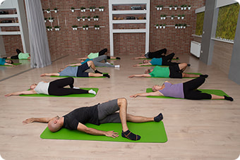 Grupa muškaraca i žena u centru za vježbanje leži na zelenim prostirkam uz ogledalo i radi s fizioterapeutom vježbe za fleksibilnost. 