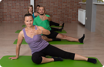 Lijepa mlada žena i muškarci iza nje na zelenim prostirkama rade vježbe medicinske gimnastike. 
