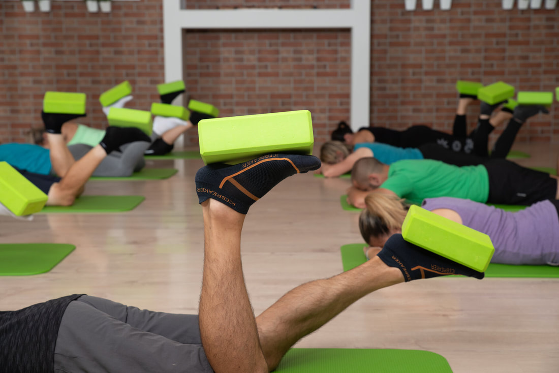 Grupa ljudi leži na trbuhu na zelenim prostirkama i fizioterapeut im pokazuje vježbe propriocepcije za stopala.